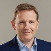 Portrait of Andrew McLean, wearing a navy blazer and posed against a neutral gray background, looking directly at the camera.