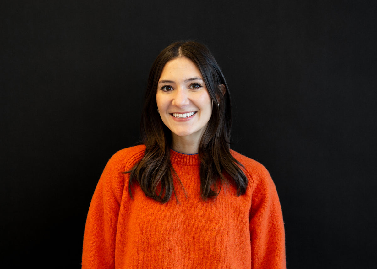 Portrait of a smiling woman with long, dark brown hair, wearing an orange sweater against a black background.