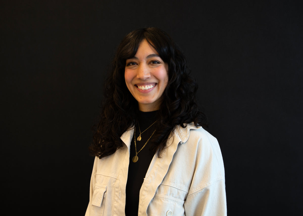 Portrait of a smiling woman with dark, curly hair, wearing a black shirt and a light-colored jacket against a black background.