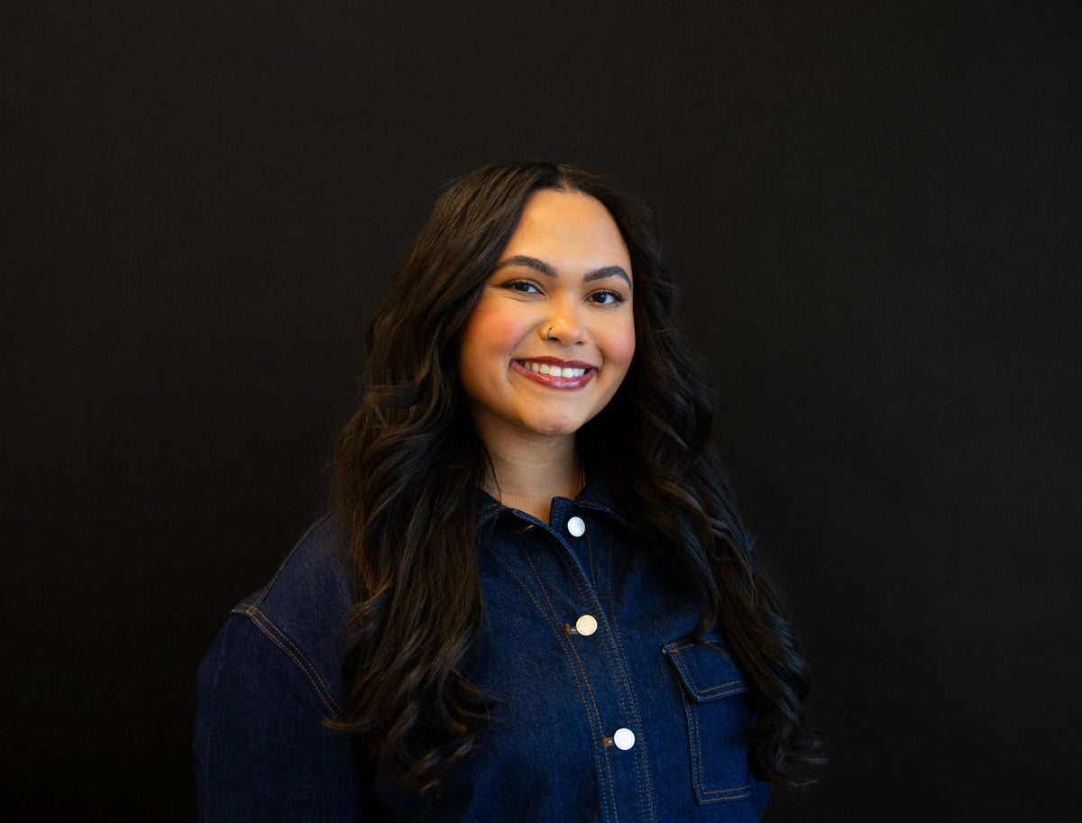 Portrait of a smiling woman with long, dark wavy hair, wearing a denim jacket against a black background.