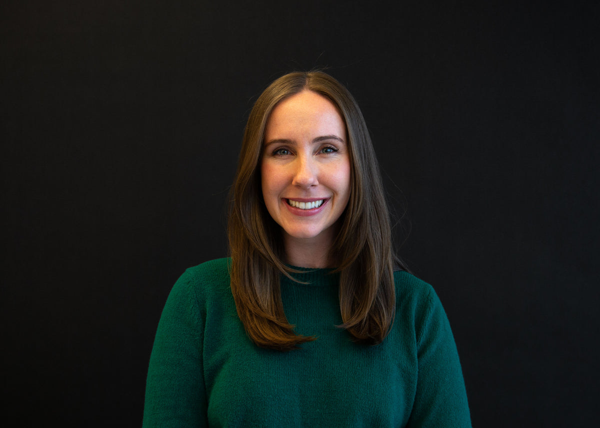 Portrait of a smiling woman with long, straight brown hair, wearing a dark green sweater against a black background.