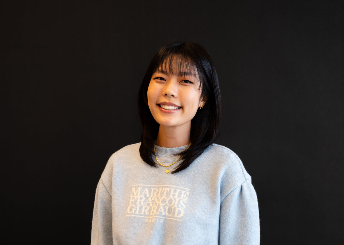 Portrait of a smiling woman with shoulder-length black hair, wearing a light blue sweatshirt with text on the front, against a black background.