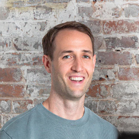 Smiling man in a muted green shirt poses in front of a rustic brick wall.