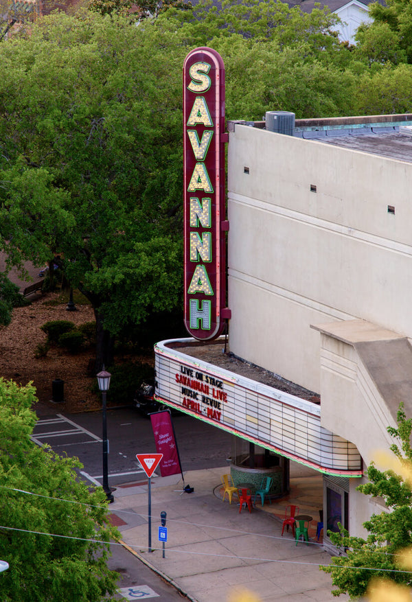 Exterior of the Savannah Theatre with a vintage vertical sign reading 'SAVANNAH' in bright lights. The marquee below advertises a live musical revue. Green trees and a sidewalk café with colorful chairs are visible in the foreground.