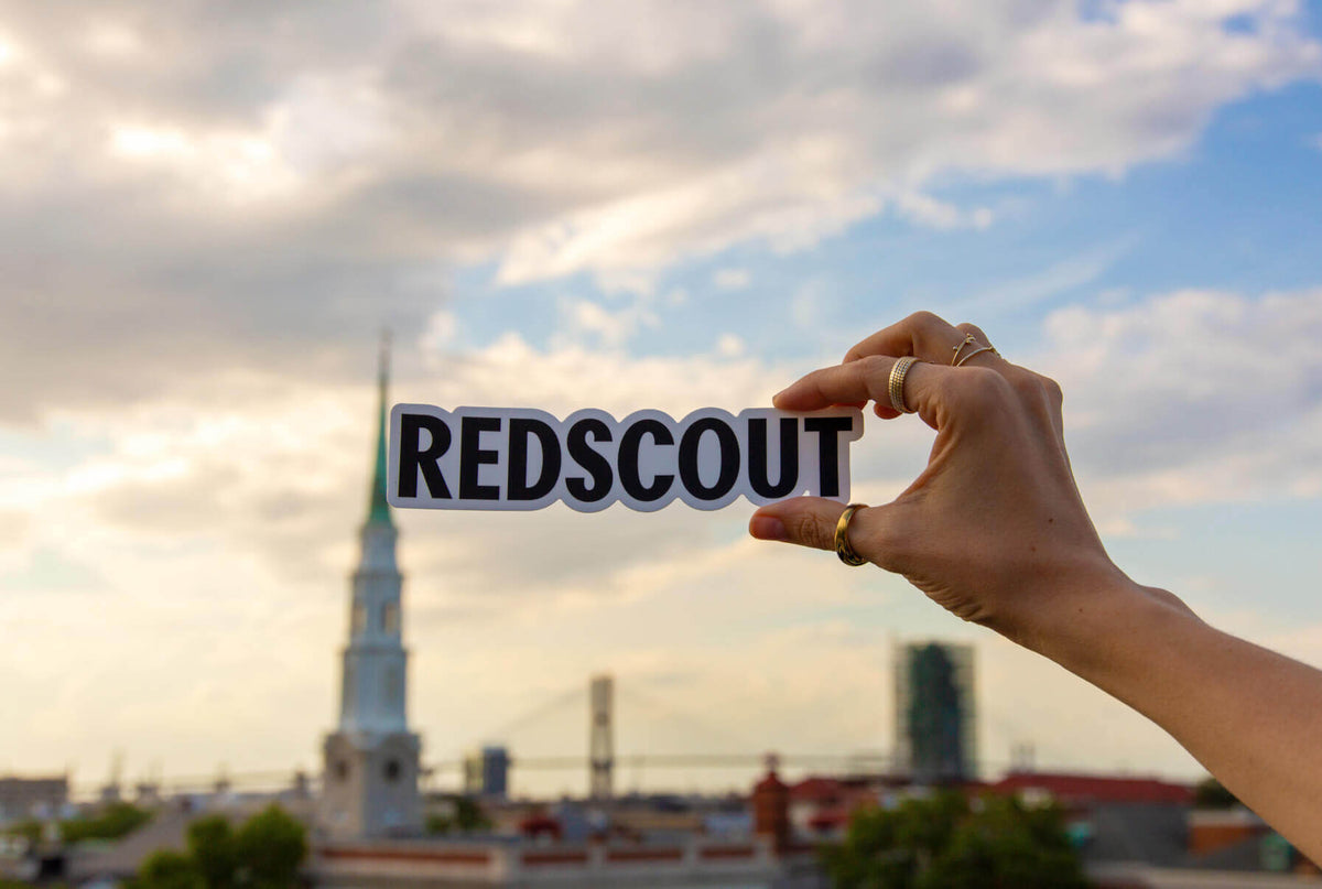 A hand holding a 'REDSCOUT' sticker against a cityscape backdrop featuring a church steeple and a suspension bridge under a partly cloudy sky.