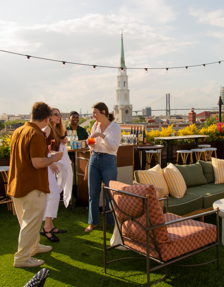 Group of people enjoying drinks and conversation on a stylish rooftop bar with string lights, overlooking a cityscape featuring a church steeple and a suspension bridge in the background.