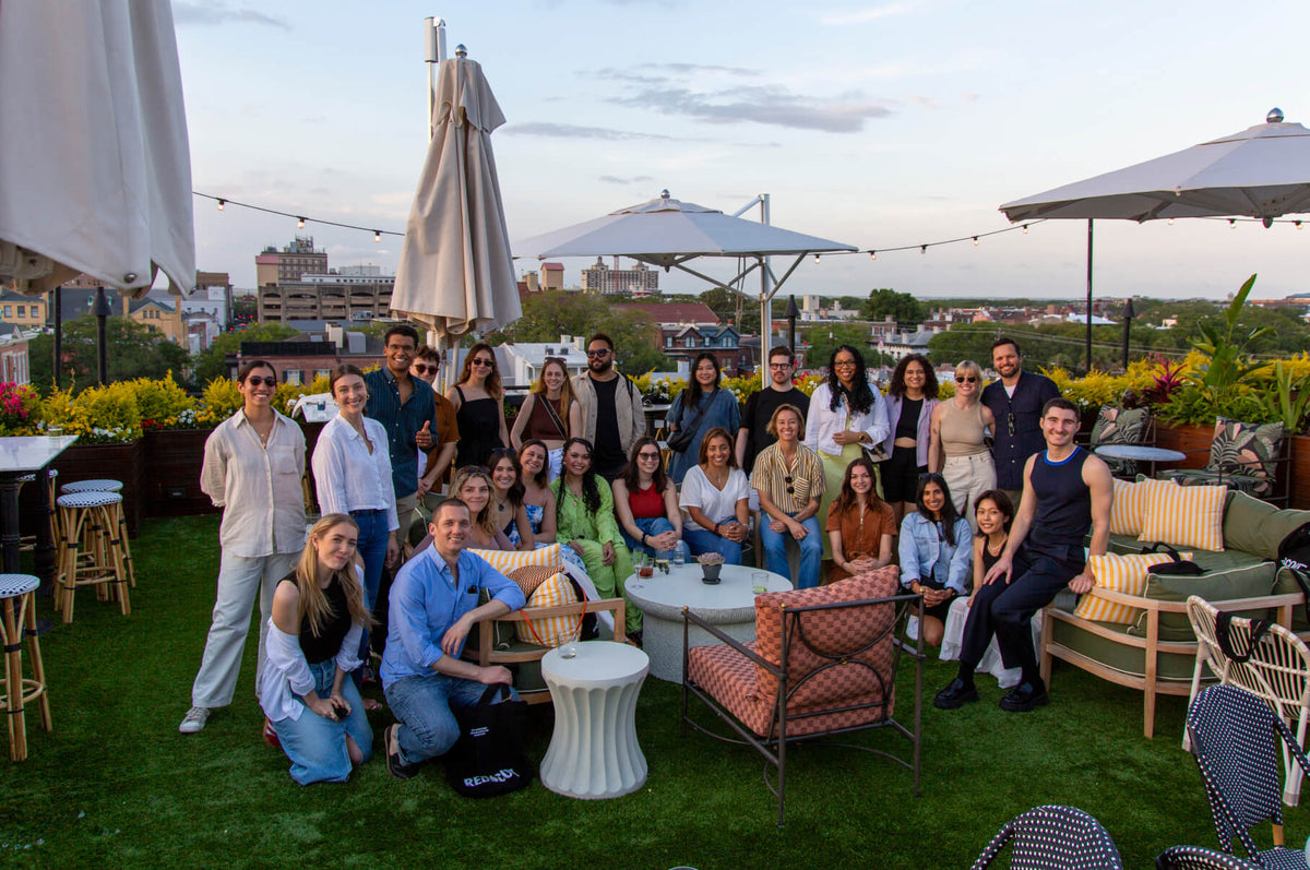 A large group of people posing together on a rooftop lounge with string lights, umbrellas, and greenery, overlooking a cityscape at sunset.